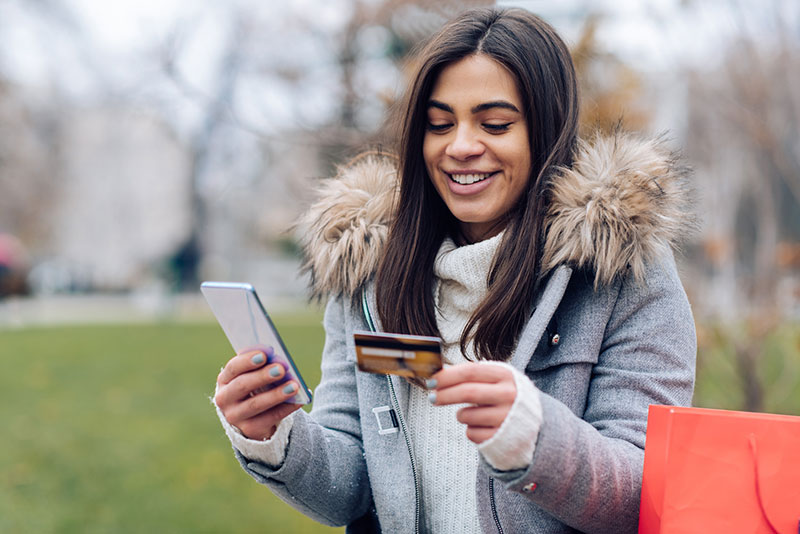happy young woman uses credit card to donate on phone outdoors